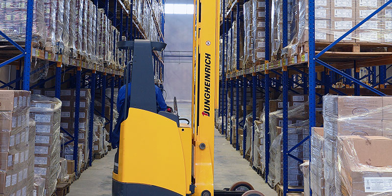 Yellow pedestrian stacker lifts boxes to put them on a pallet rack at the huge warehouse.
